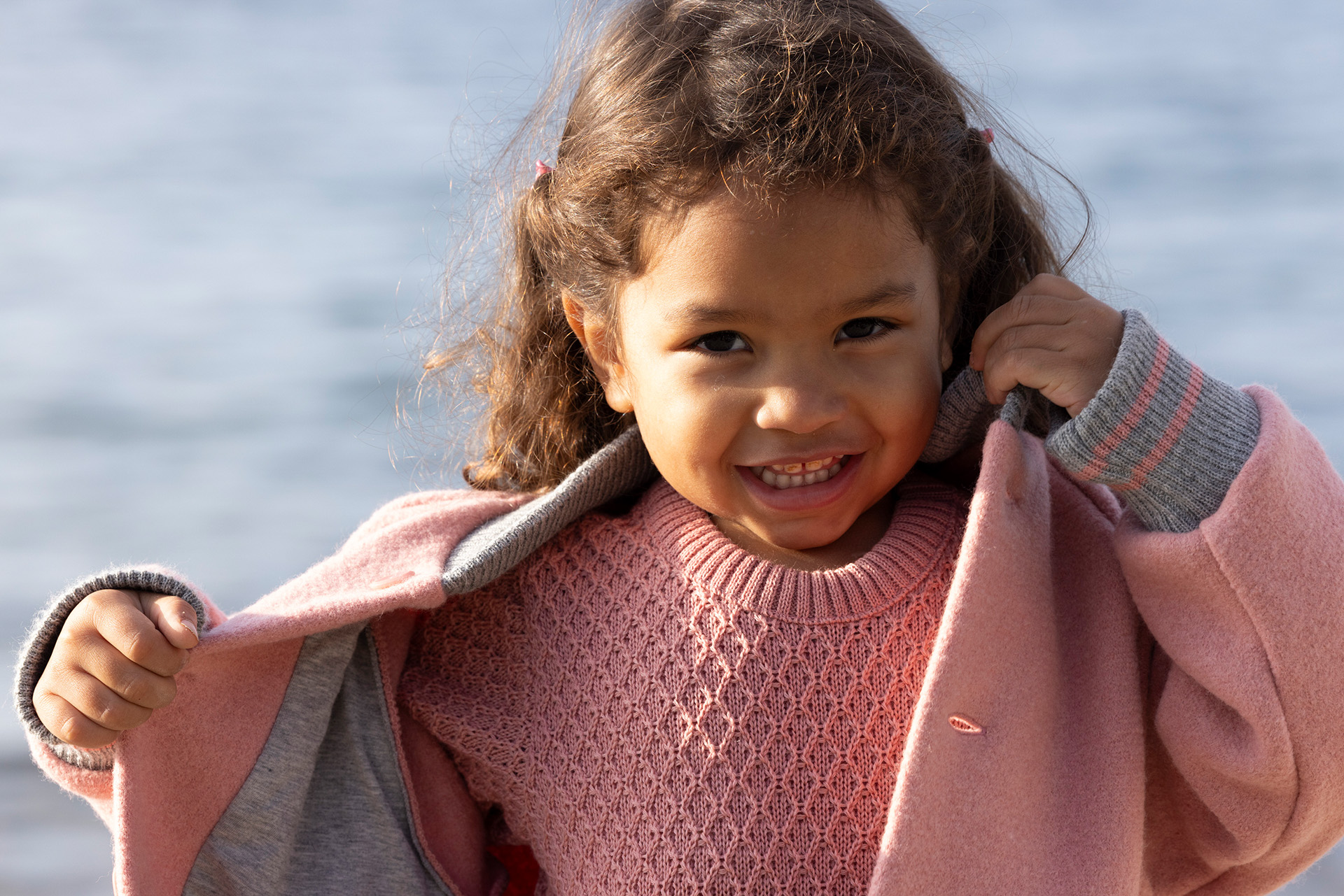 Lächelndes Mädchen mit hellbrauner Haut in disana College-Jacke Softpink mit passendem Aran-Pullover vor Wasserhintergrund