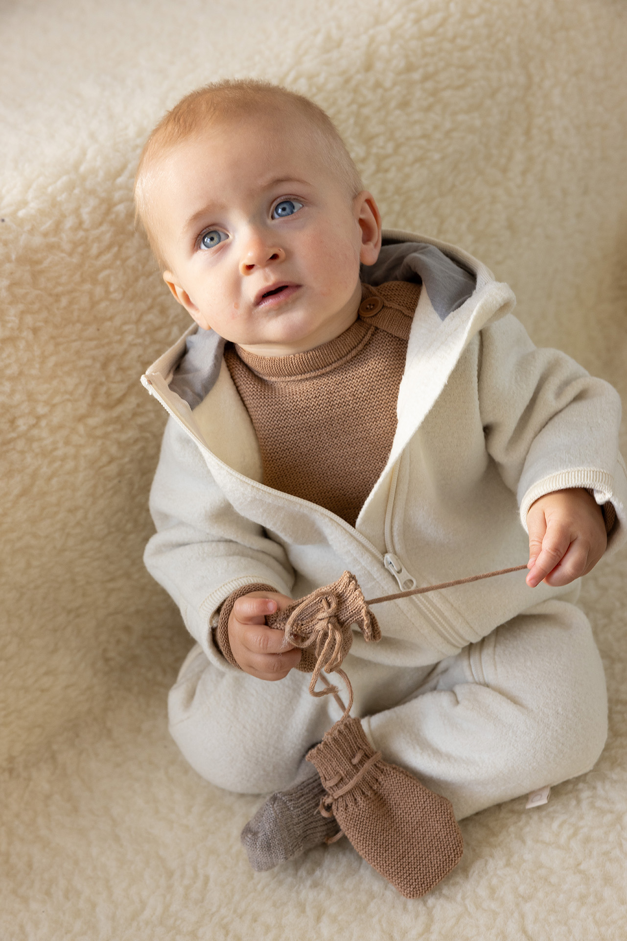Blauäugiges Baby in disana Zipp-Overall Natur, Baby-Pullover & Strick-Handschuhe in Karamell auf hellem Lammfell-Hintergrund