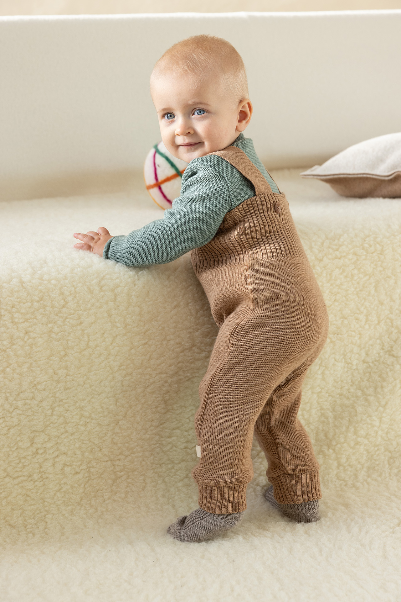 Blondes Baby in disana Strick-Trägerhose Karamell, Melange-Pullover Jade aus Bio-Merinowolle auf flauschigem Lammfell stehend