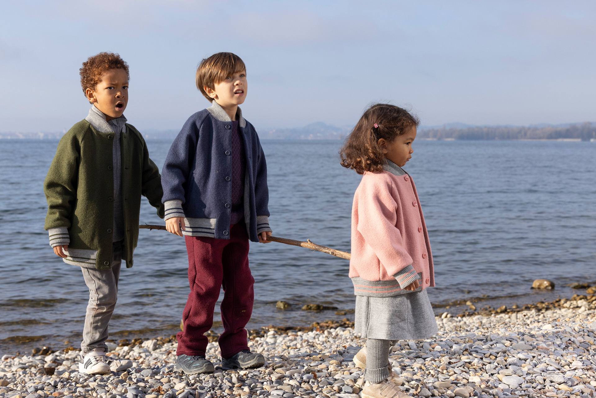 3 Kinder am Kiesstrand am Seeufer in disana College-Jacken Oliv, Indigo, Softpink halten einen horizontalen Holzstock.