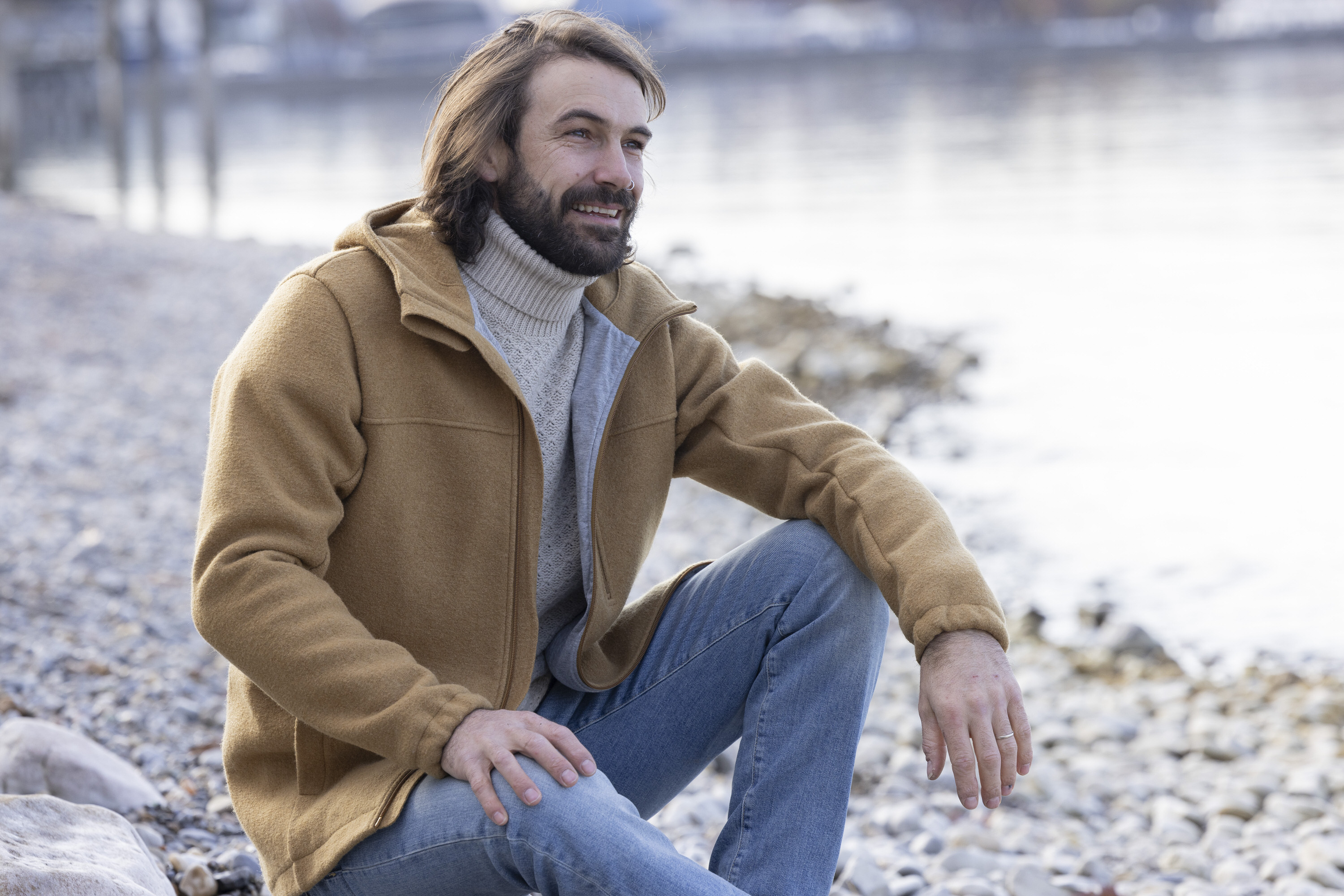 Lächelnder Mann mit dunkelbraunen Haaren und Bart in disana Herren-Jacke Toffee und Jeanshose sitzend an einem Seeufer