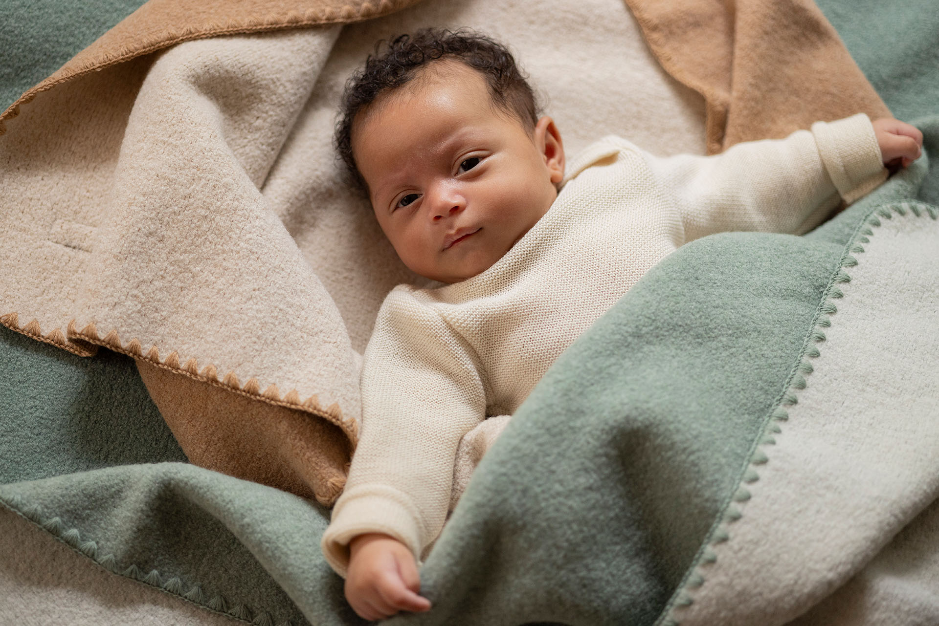 Baby mit hellbraunem Hautton eingekuschelt in disana Doubleface-Babydecken in den Farben Karamell-Natur und Jade-Natur