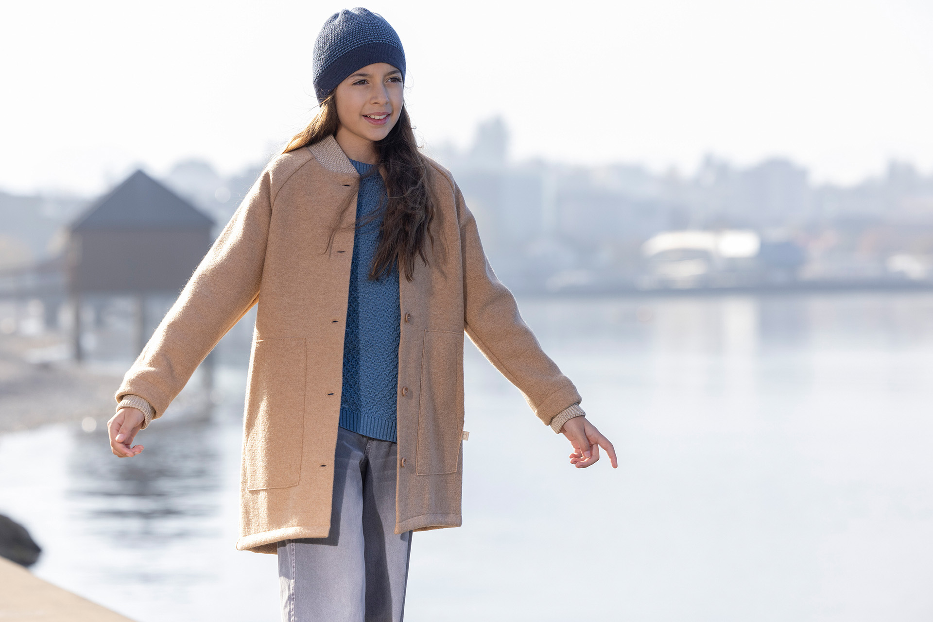 Mädchen mit Beanie Indigo-Taubenblau, lange braune Haare im disana Walk-Mantel, Karamell balanciert an einer Uferpromenade