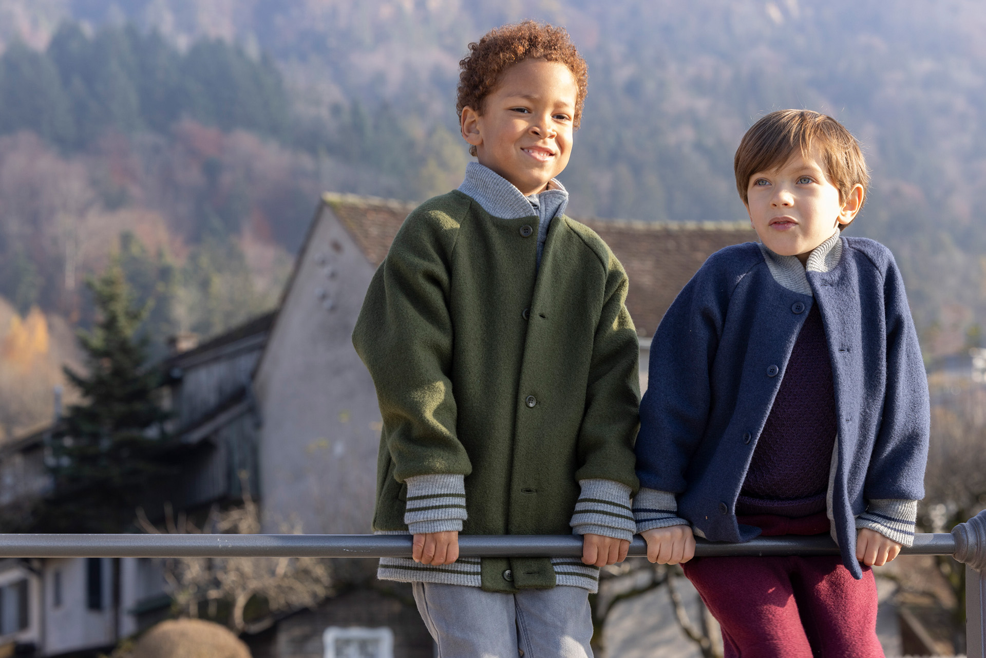 2 Kinder in disana College-Jacken, Oliv & Indigo mit gestreiften Strickbündchen an Kragen & Ärmeln vor Bergkulisse