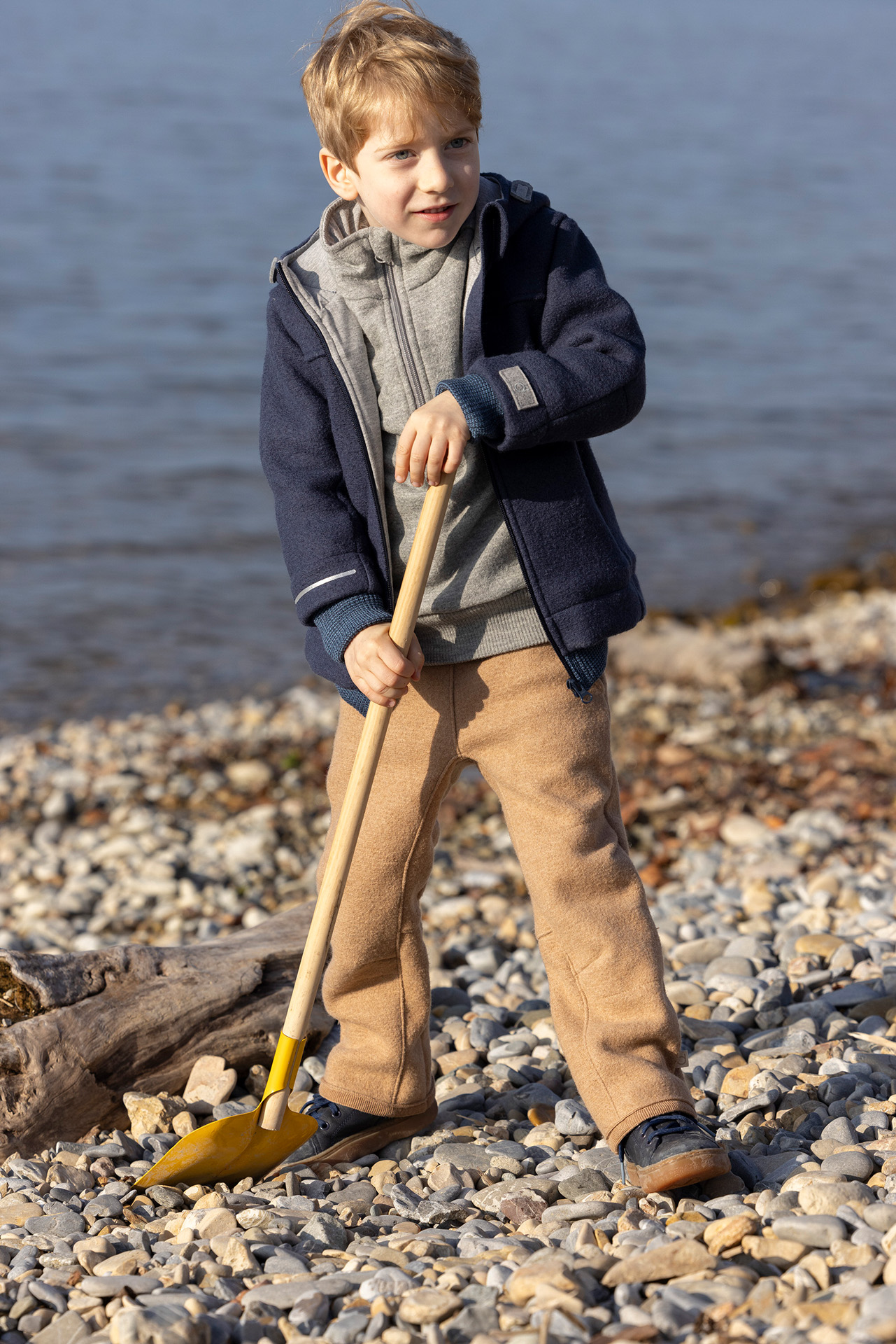 Blondes Kind in disana Outdoor-Jacke Indigo, Walk-Hose Karamell & Troyer Grau hält eine Schaufel am Kiesufer eines Sees