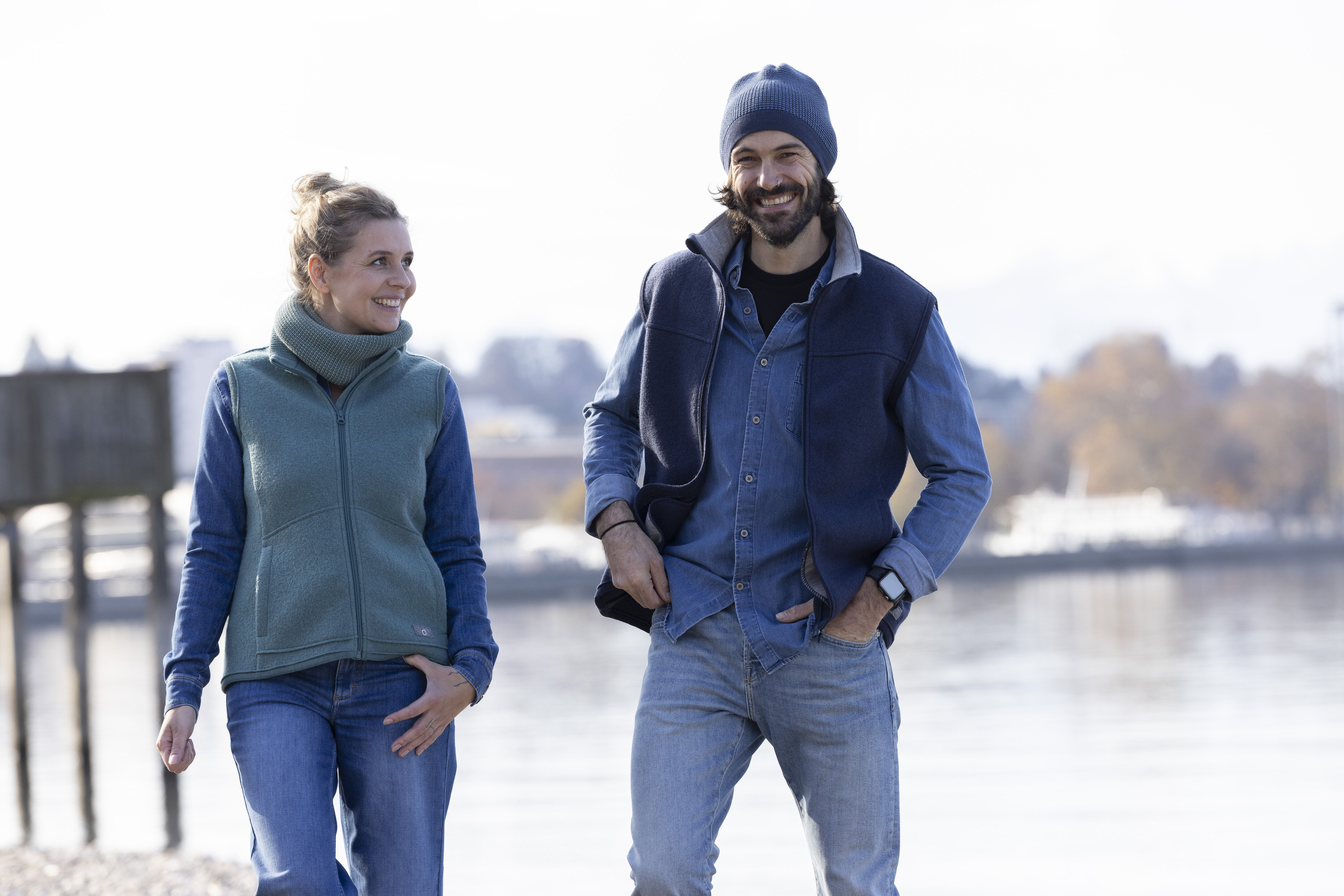 Mann und Frau spazieren an einem Seeufer in disana Damen-Weste in Eukalyptus und Herren-Weste in Indigo mit Beanie