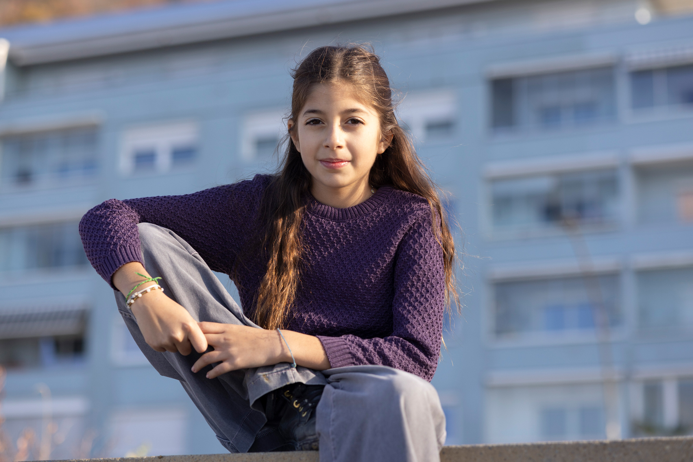 Porträt eines Mädchens mit langen braunen Haaren in disana Aran-Pullover Aubergine und grauer Hose vor blauem Gebäude