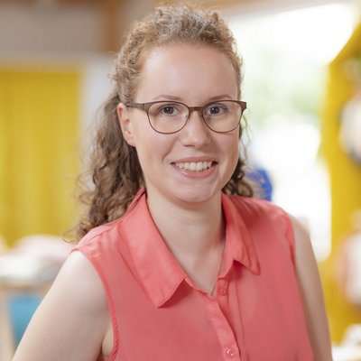 Doreen Böbel, Verwaltungsangestellte bei disana in oranger Bluse vor unscharfem Hintergrund lächelt in die Kamera.