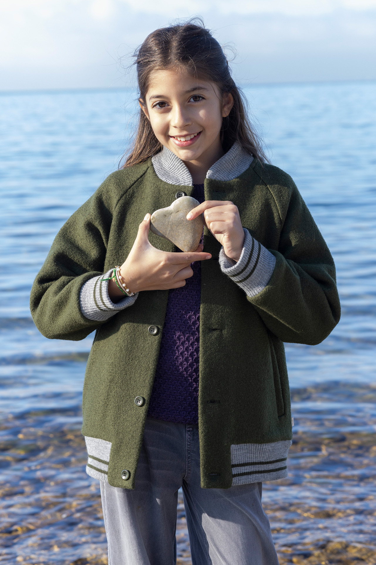 Mädchen mit braunen Haaren in College-Jacke, Oliv & Aran-Pullover Aubergine hält einen Stein in Herzform am Seeufer